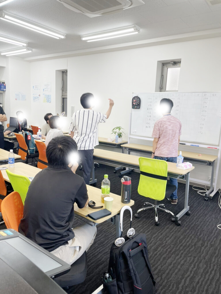 ダーツ大会の様子 ホワイトボードに吊るした的にダーツを投げる人、それを見守る参加者たち。